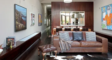 Living room with brown leather sofa, striped cushions, artwork on walls, and a dining area in the background.
