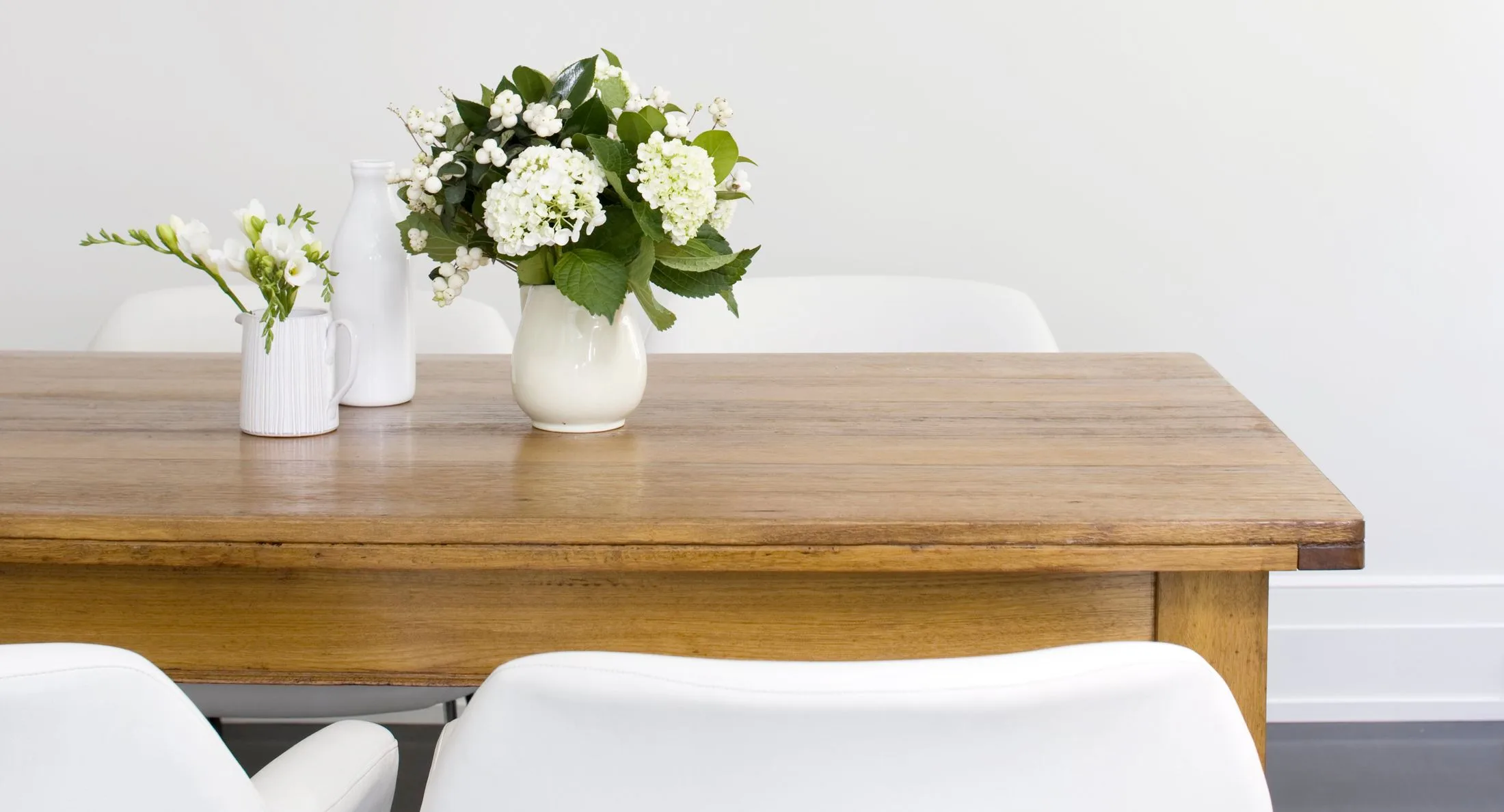 A wooden table with two vases of white flowers against a plain wall.