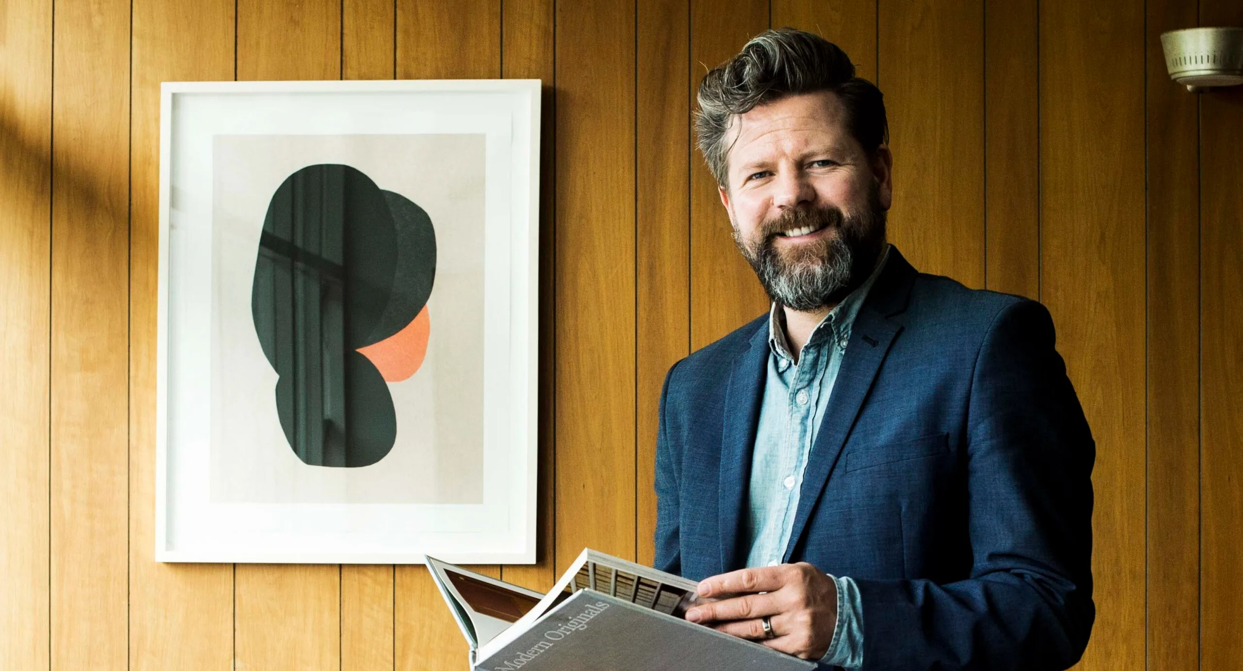 Man in a suit holding a book, smiling, with abstract art on a wood-paneled wall background.