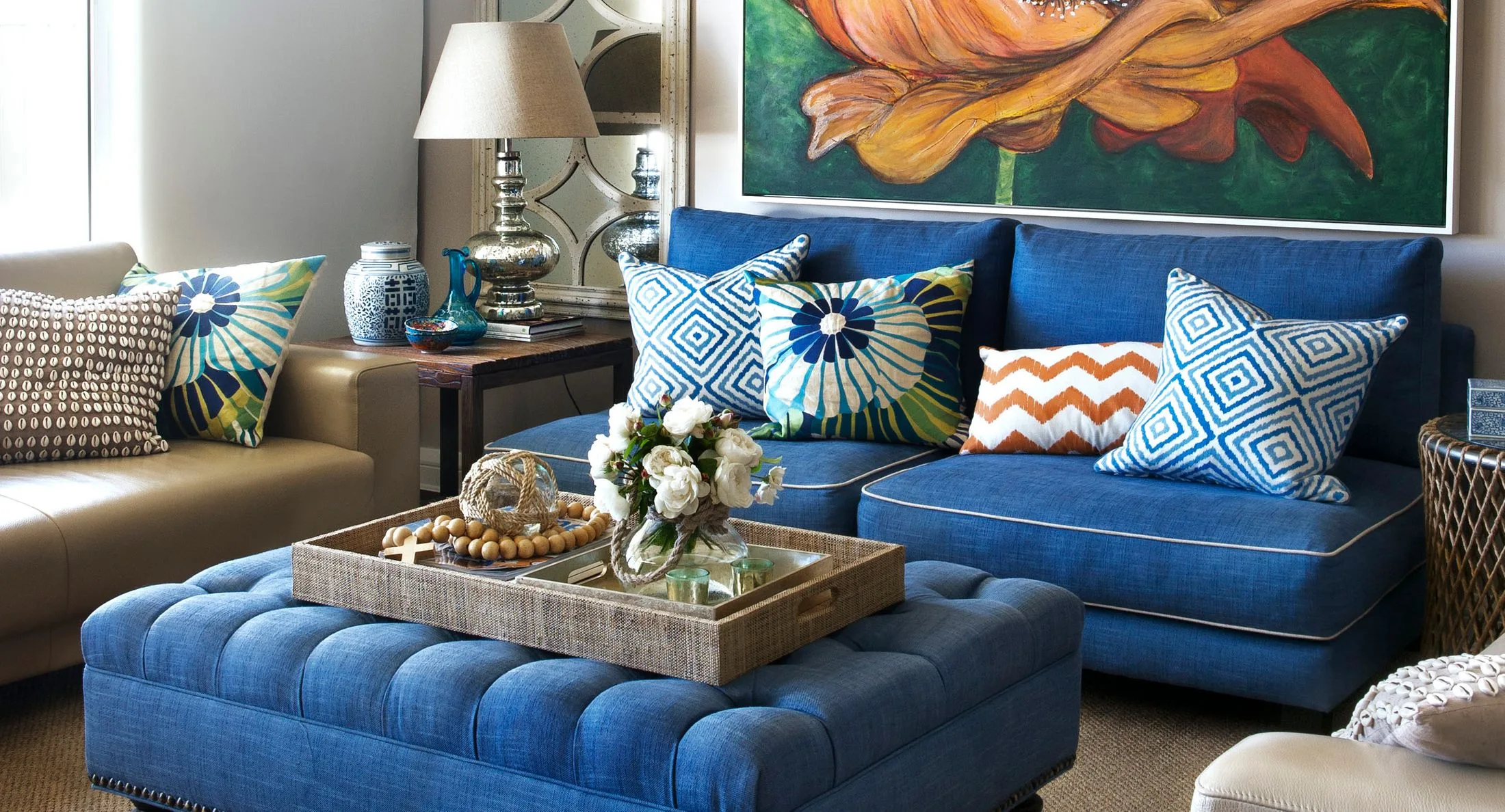 Living room with blue sofa, colorful cushions, large floral painting, and a tray with flowers on an ottoman.