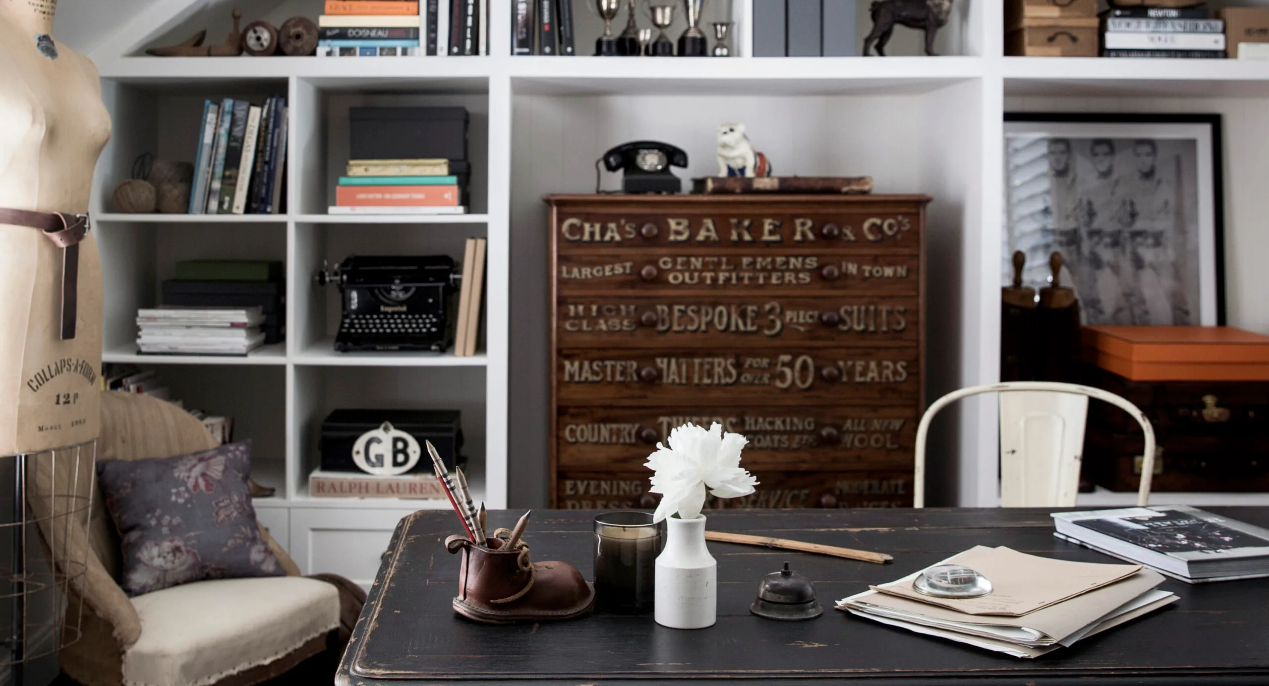 Office with vintage decor, typewriter, mannequin, shelves of books, and a desk with flowers and writing tools.