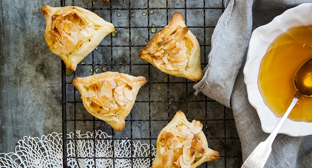 Four almond-topped hamantaschen on a cooling rack with a bowl of honey and spoon beside them.