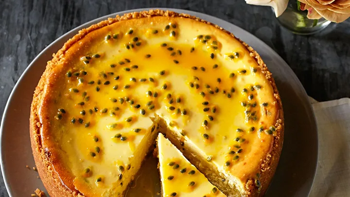 A round passionfruit cheesecake with a slice cut, topped with seeds, beside yellow roses in a vase.