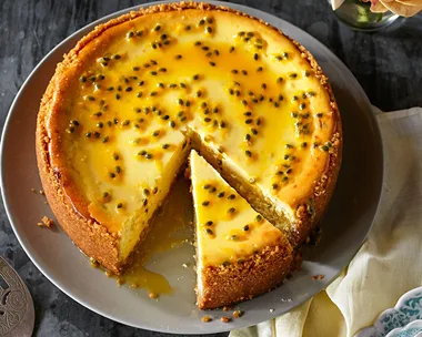 A round passionfruit cheesecake with a slice cut, topped with seeds, beside yellow roses in a vase.
