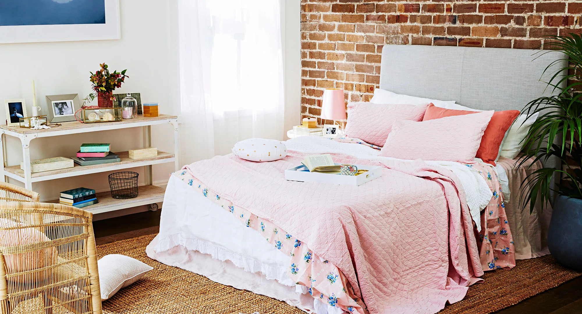 Cozy bedroom with a pink quilt, brick wall, plant, wicker chair, and shelf with decor under a bright window.