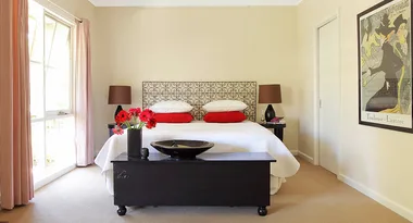 Cozy bedroom with a patterned headboard, red pillows, and a painting on the wall. Sunlight streams through a large window.