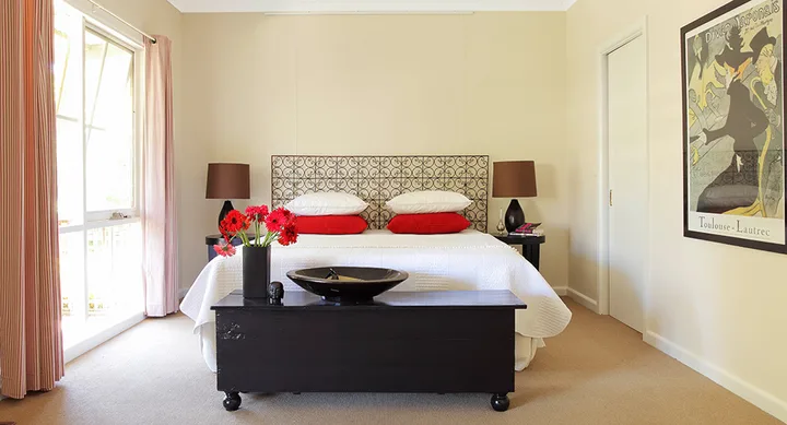 Cozy bedroom with a patterned headboard, red pillows, and a painting on the wall. Sunlight streams through a large window.