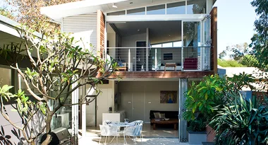 Modern two-story house with large windows, balcony, and patio surrounded by greenery.