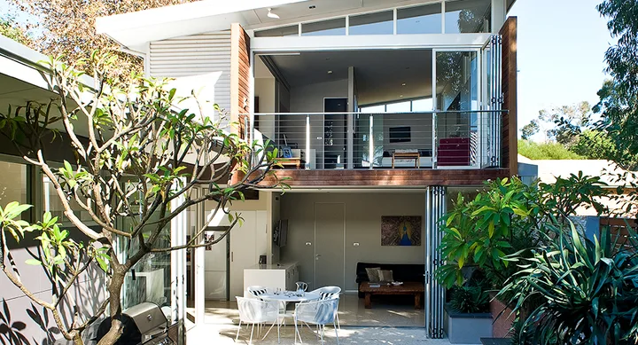Modern two-story house with large windows, balcony, and patio surrounded by greenery.