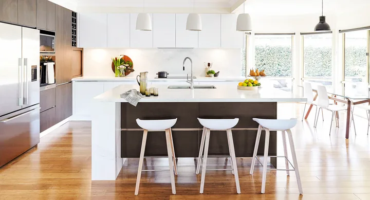 Modern kitchen with wooden floors, large island, white stools, pendant lights, and a view of greenery through windows.