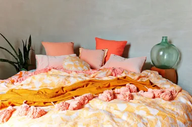 Cozy bed with pink and orange pillows, patterned duvet, orange throw, plant, and large glass vase on nightstand.