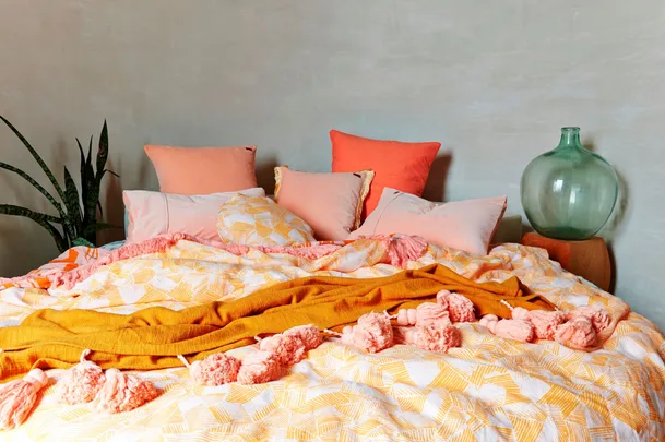 Cozy bed with pink and orange pillows, patterned duvet, orange throw, plant, and large glass vase on nightstand.