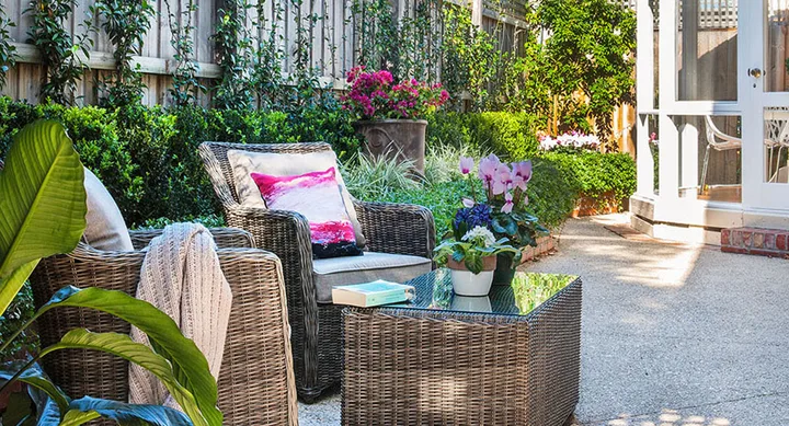 Outdoor patio with wicker chairs, cushion, blanket, plants on a table, and lush greenery surrounding the area.