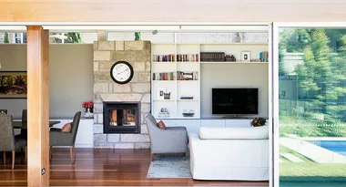 Living room with fireplace, clock, bookshelves, TV, armchairs, and view of a pool through a glass door.
