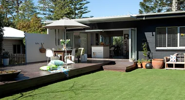 Outdoor patio with umbrella, chairs, and table set on wooden deck attached to a modern house, overlooking a garden.