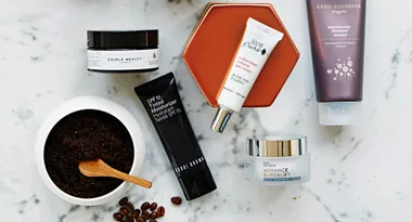 Assorted skincare products and ground coffee in a bowl with a wooden spoon on a marble surface.