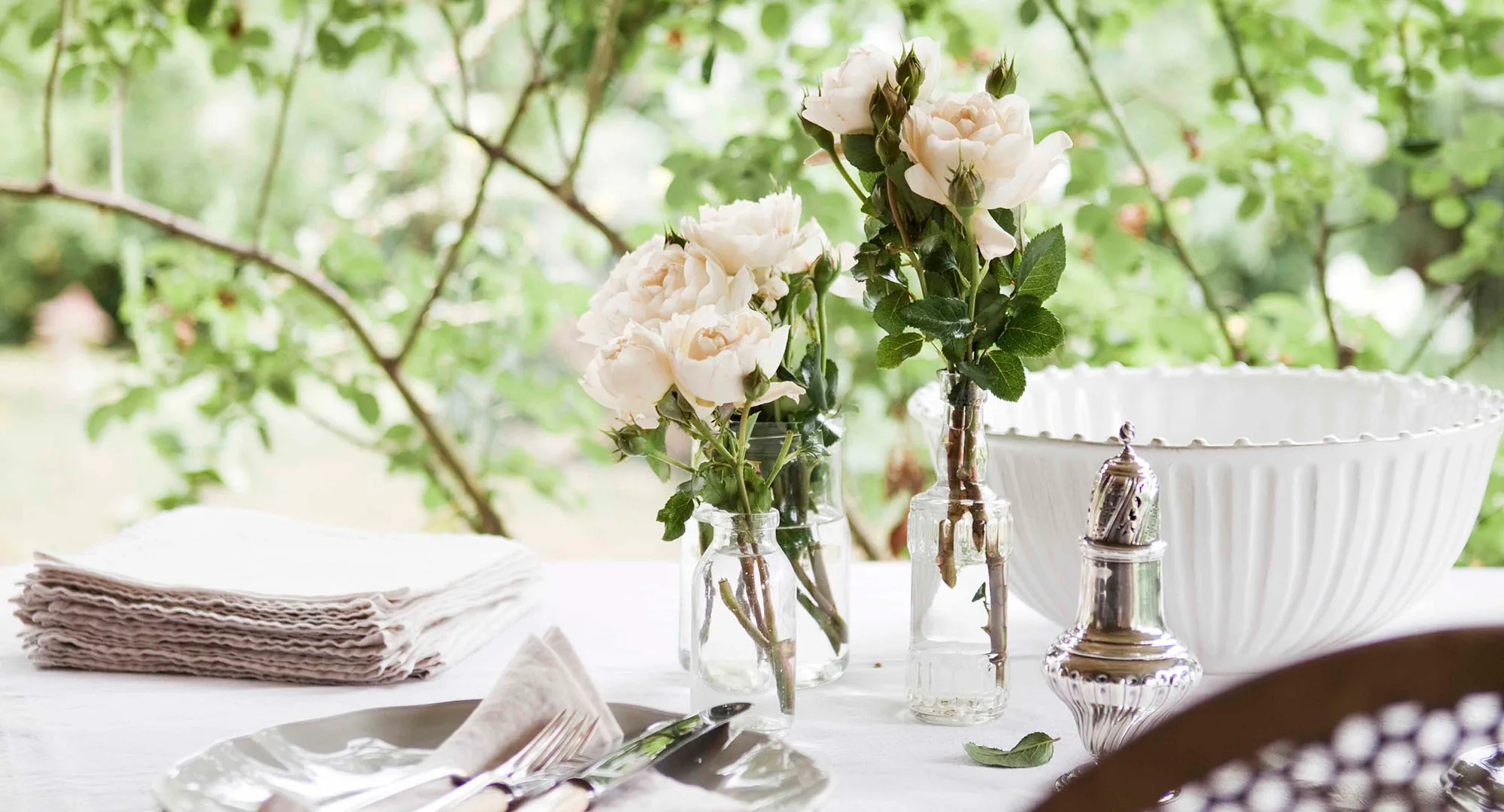 White roses in glass vases on a table with napkins, cutlery, and a white bowl in a garden setting.