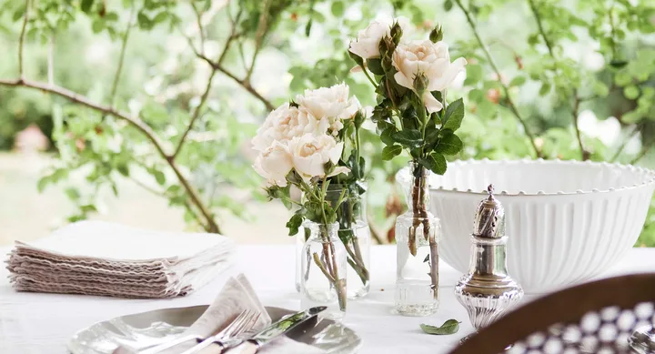 White roses in glass vases on a table with napkins, cutlery, and a white bowl in a garden setting.
