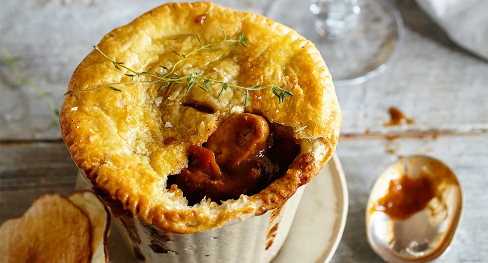 Golden puff pastry pie with meat filling, garnished with thyme, served on a rustic plate with a spoon beside it.