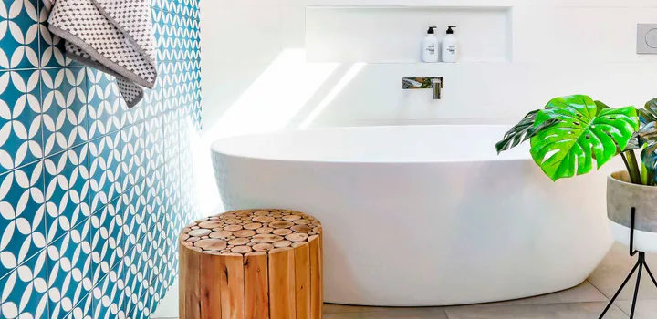 Luxury bathroom with a freestanding tub, wooden stool, plant, geometric blue tiles, and sunlight streaming.