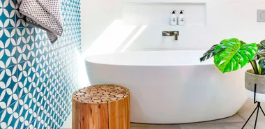 Luxury bathroom with a freestanding tub, wooden stool, plant, geometric blue tiles, and sunlight streaming.
