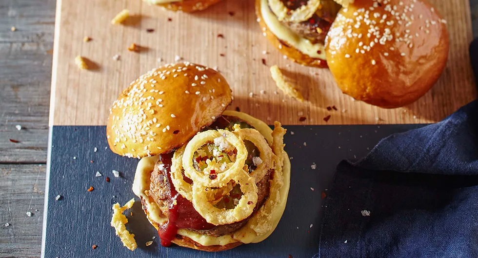 Burger with sesame seed bun, topped with cheese, fried onion rings, and sauce, on a wooden board.