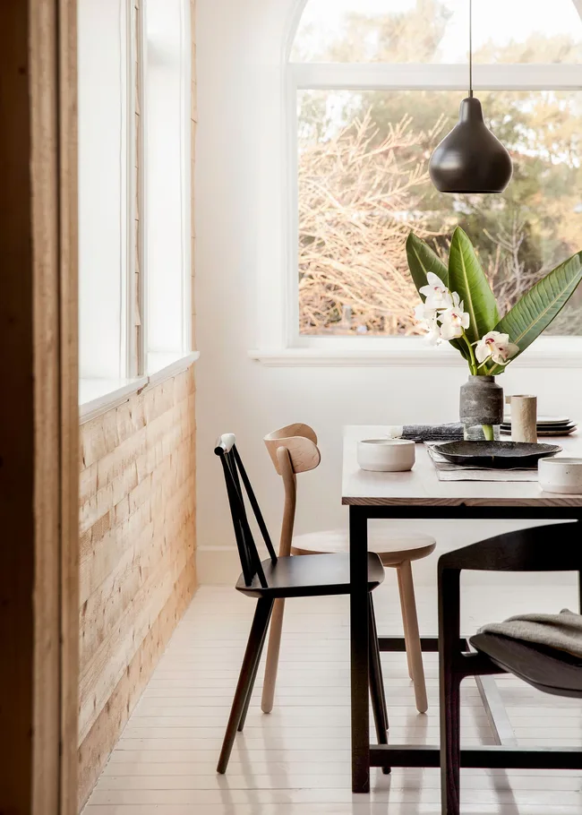 Minimalist dining area with a wooden table, black chairs, a pendant light, and a vase with white flowers.