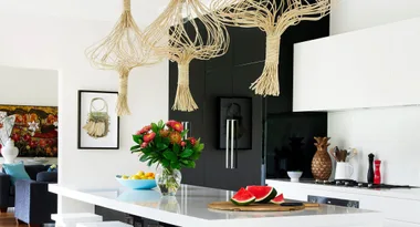 Kitchen with modern rattan light fixtures, white countertops, floral arrangement, and sliced watermelon on a board.