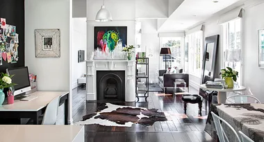 Modern, bright open-plan living room with a fireplace, artwork, dark wood floors, and a study nook with a computer.