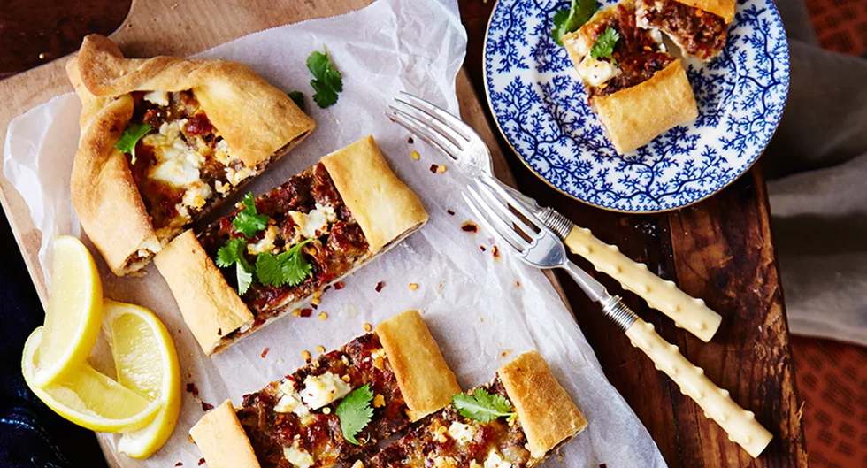 Turkish pide with meat and cheese garnished with herbs, served on a wooden board, accompanied by lemon wedges and cutlery.