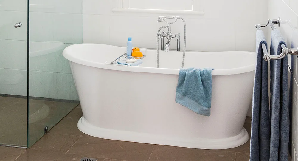 A white freestanding bathtub with a chrome faucet, blue towel, rubber duck, and toiletries, next to a glass shower.