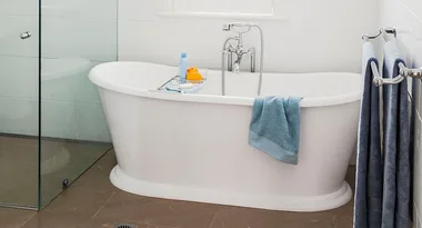 A white freestanding bathtub with a chrome faucet, blue towel, rubber duck, and toiletries, next to a glass shower.