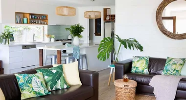 Open-plan living room with brown leather sofas, green leaf-patterned pillows, kitchen area, and large round wall mirror.