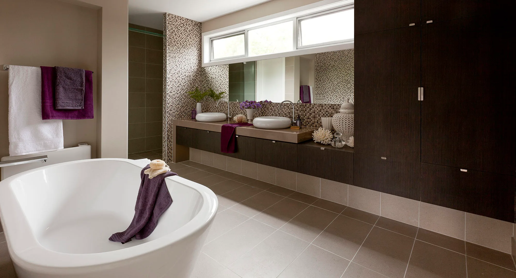 Modern bathroom with white tub, purple towels, dual sinks, and mosaic tiles under large windows.