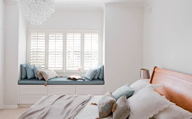 Cozy bedroom with chandelier, window seat, blue cushions, and a neatly made bed.