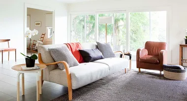 Bright living room with a beige sofa, colorful cushions, leather chair, and large window with garden view.