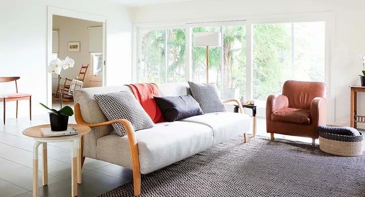 Bright living room with a beige sofa, colorful cushions, leather chair, and large window with garden view.