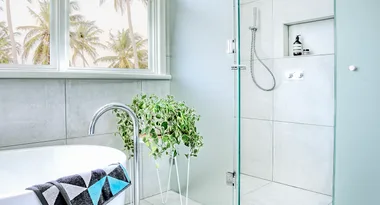 Modern bathroom with a white tub, green plant, palm trees outside the window, and a sleek shower area with toiletries.