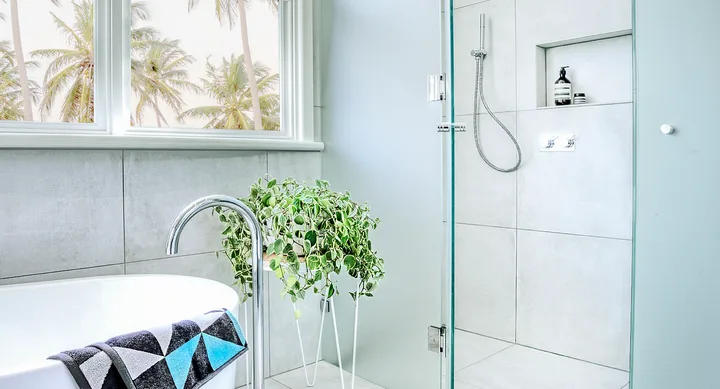 Modern bathroom with a white tub, green plant, palm trees outside the window, and a sleek shower area with toiletries.