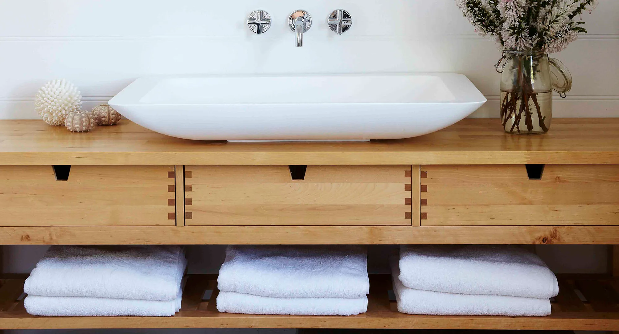 Modern bathroom vanity with a white sink, wooden drawers, decorative shells, and a jar of flowers.