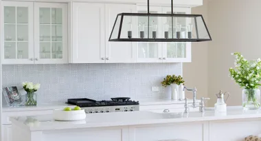 Bright modern kitchen with white cabinets, patterned backsplash, flowers, and island with decorative lighting above.