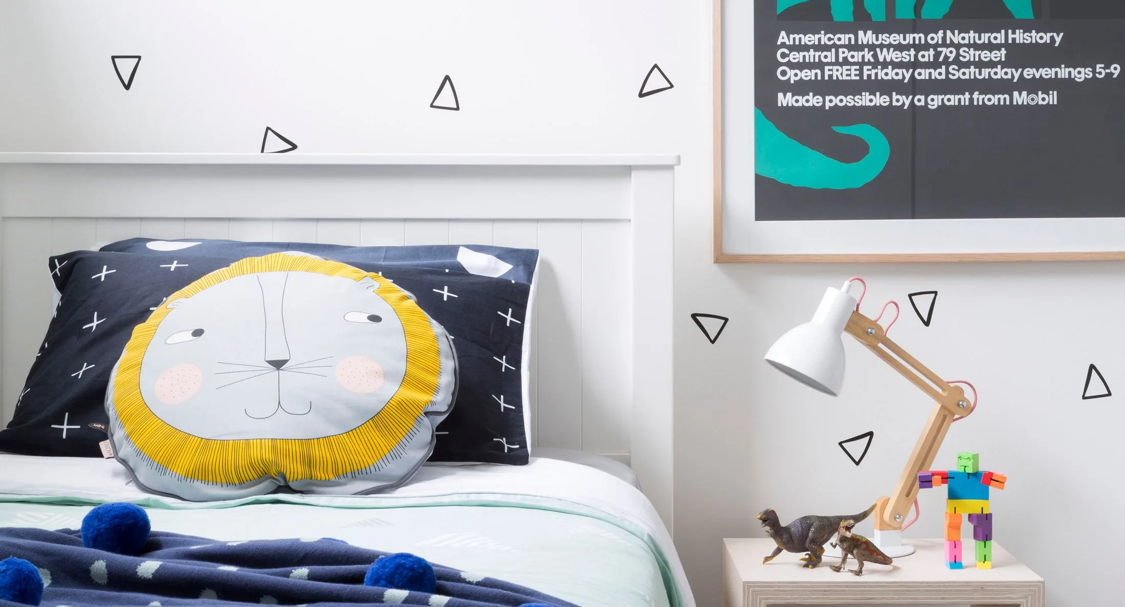 Cozy kids' room with lion face pillow, geometric decor, and a side table with lamp, toys, and framed museum poster.