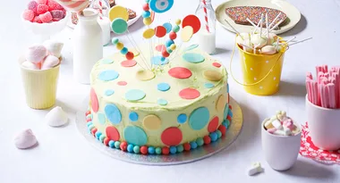 Colorful polka dot cake with candy decorations on a table surrounded by sweets and drinks.
