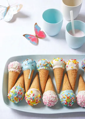 Colorful ice cream cones with sprinkles on a tray, surrounded by cups and butterfly decorations on a white table.
