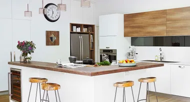 Modern kitchen with large island, wooden stools, pendant lights, clock, and stainless steel appliances.