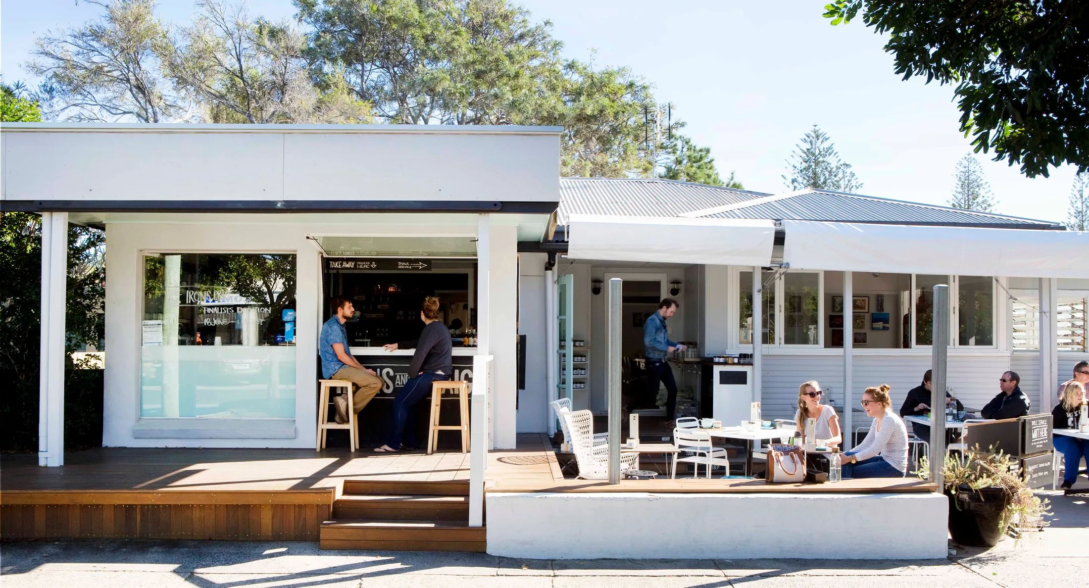 Outdoor cafe with people sitting at tables under a clear blue sky, trees surround the modern white building.