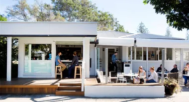 Outdoor cafe with people sitting at tables under a clear blue sky, trees surround the modern white building.