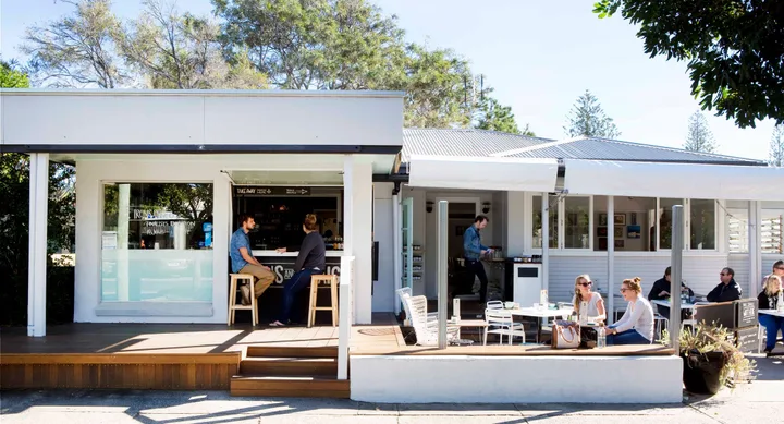 Outdoor cafe with people sitting at tables under a clear blue sky, trees surround the modern white building.