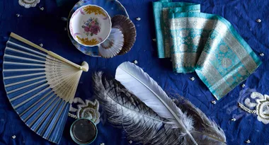 A collection of vintage items including a fan, teacup, napkin, and feathers on a blue fabric background.