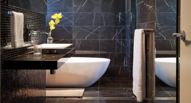 Modern bathroom with black tiles, white bathtub, floating sink, and yellow flowers in a vase.